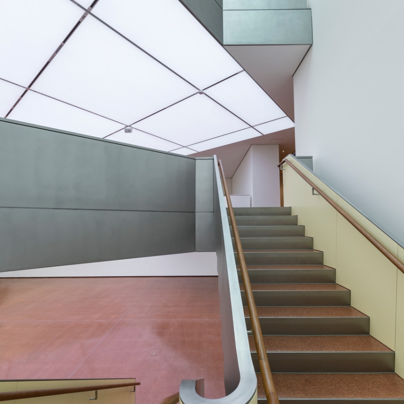 Modern staircase with a wooden railing, leading to a spacious gallery with minimalist design and soft, diffused lighting.