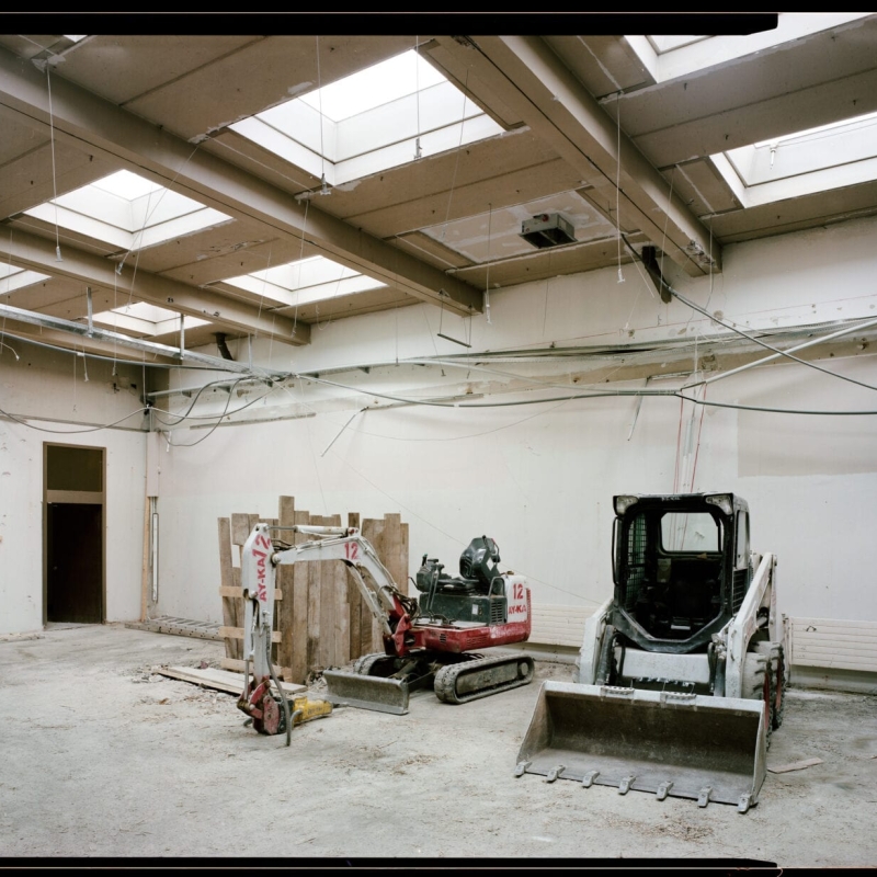 Empty construction site with two small excavators, wooden pallets, and exposed ceiling beams with skylights.
