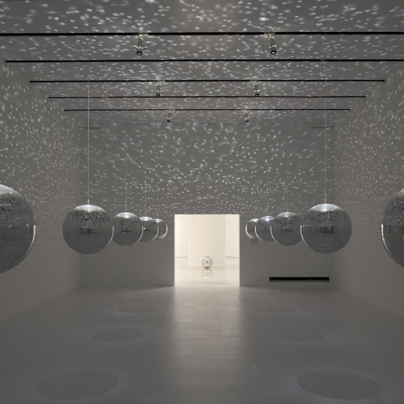 Helle Galerie mit hängenden Discokugeln, die Lichtmuster an die Wände werfen. Ein Durchgang führt in einen weiteren Raum.