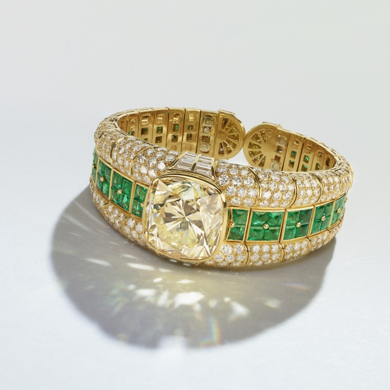 Gold bracelet featuring a large yellow diamond centerpiece, surrounded by sparkling white diamonds and green gemstones.