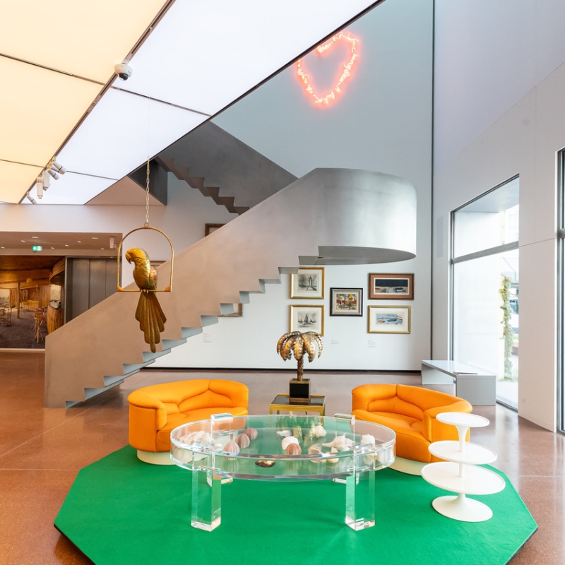 Moderne Lounge mit orangefarbenen Sesseln, gläsernem Tisch, grüner Teppich und einer Treppe im Hintergrund.