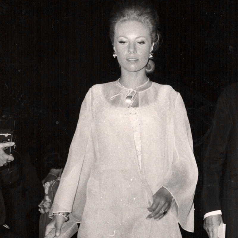 A woman in a flowing, light-colored gown walks gracefully, holding a clutch, with a soft expression and elegant hairstyle.