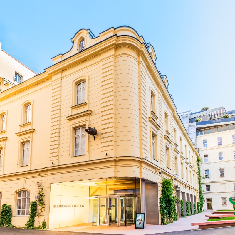 The picture shows the building of the Heidi Horten Collection.
It is located in Hanuschhof, in the center of Vienna.
The building is bright yellow and has a neoclassical façade with large windows.
The shape is reminiscent of an old city palace, but has been renovated in a modern style.
There is a large glass door at the entrance.
Next to it is written in silver lettering: “Heidi Horten Collection”.
A digital screen displays information about the museum.
On the right of the picture you can see a small garden with sculptures.
A large green figure is by the artist Keith Haring.
The photo shows the museum in fine weather.
The sky is blue and the light makes the place friendly and inviting.