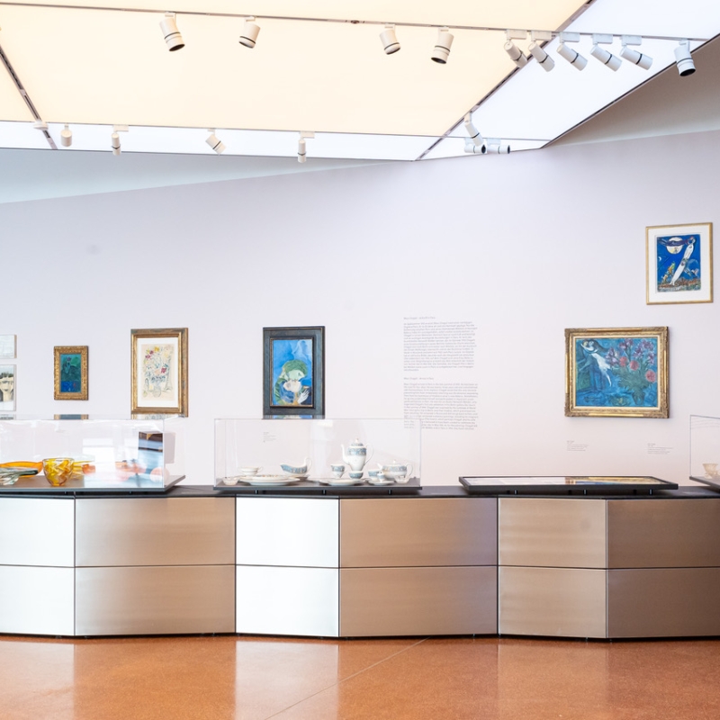 Moderne Galerie mit Gemälden an der Wand und einem Ausstellungsstück aus Glas und Metall in der Mitte.