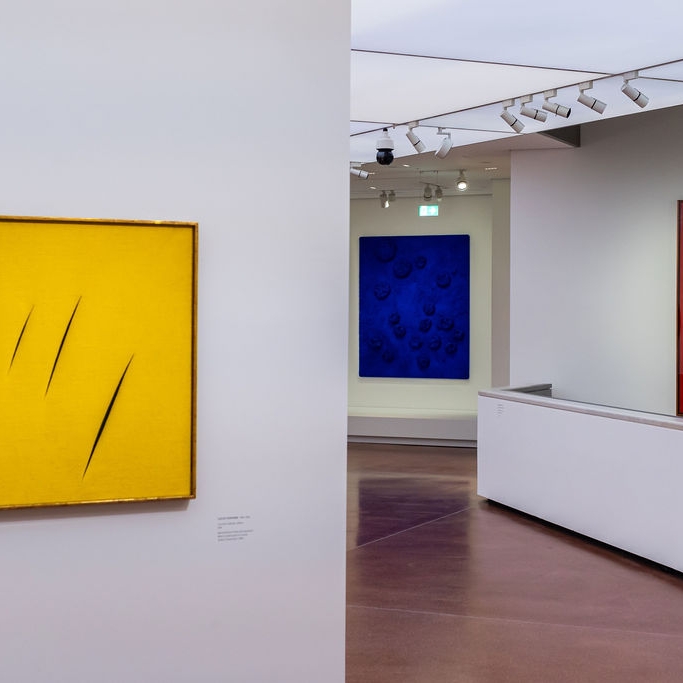 Gelbes Kunstwerk mit Einschnitten, daneben blaue und rote Gemälde in einem modernen Ausstellungsraum.