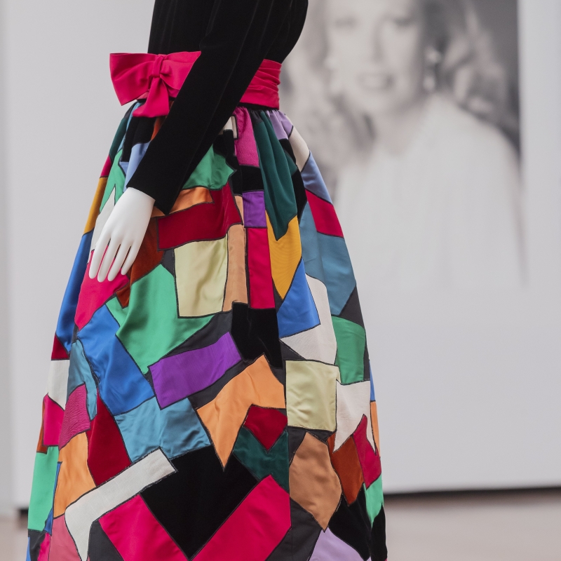 A colorful patchwork skirt with a black velvet top and a pink bow, displayed on a mannequin in an art gallery.