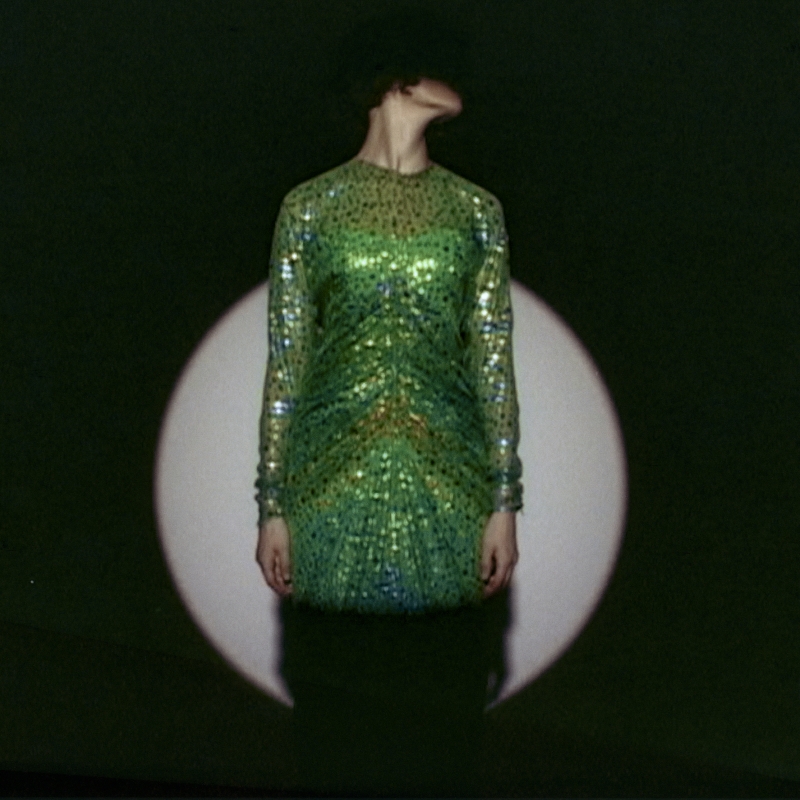 Auf dem Bild sieht man eine Person in einem grünen Kleid.
Das Kleid ist glänzend und mit vielen Pailletten oder Glitzersteinen verziert.
Die Person steht vor einem dunklen Hintergrund,
in dem ein runder, heller Lichtkreis, wahrscheinlich von einem Scheinwerfer, zu sehen ist
Der Kopf der Person ist nach links geneigt,
sodass man das Gesicht nicht sehen kann.
Das macht das Bild geheimnisvoll und spannend.