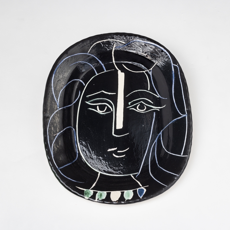 Schwarze Keramikplatte mit stilisiertem Gesicht, weißen Linien und bunten Akzenten im unteren Bereich.