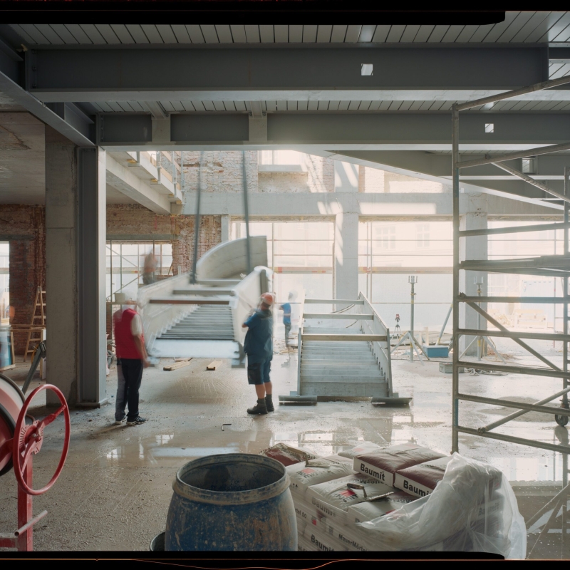 Bauarbeiten in einem großen Raum, Arbeiter heben Treppenstufen, Baustellenmaterialien und Gerüste sind sichtbar.