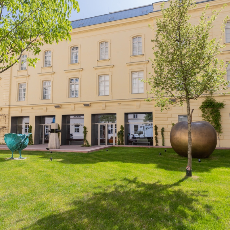 Gelbes Gebäude mit großen Fenstern, umgeben von Rasen, Bäumen und modernen Skulpturen.