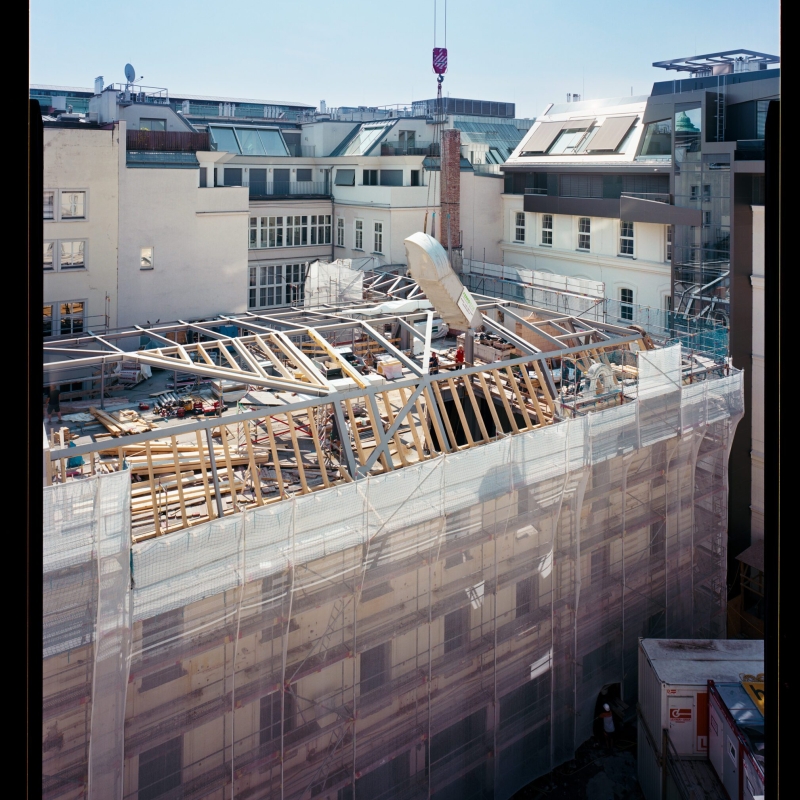 Blick auf eine Baustelle mit freiliegendem Dachstuhl, umgeben von Gerüst und Baucontainern, bei klarem Himmel.